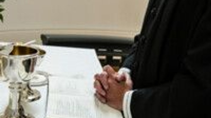 En person i religiøst tøj læser fra en åben bog under en ceremoni med en kalk i nærheden.