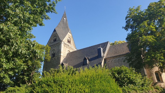Kirchturm mit spitzem Dach durch Bäume und Büsche hindurch sichtbar