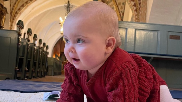 Nysgerrig baby i rød bodystocking, der kravler på en hvid overflade i et prægtigt indendørs rum med lysekroner.