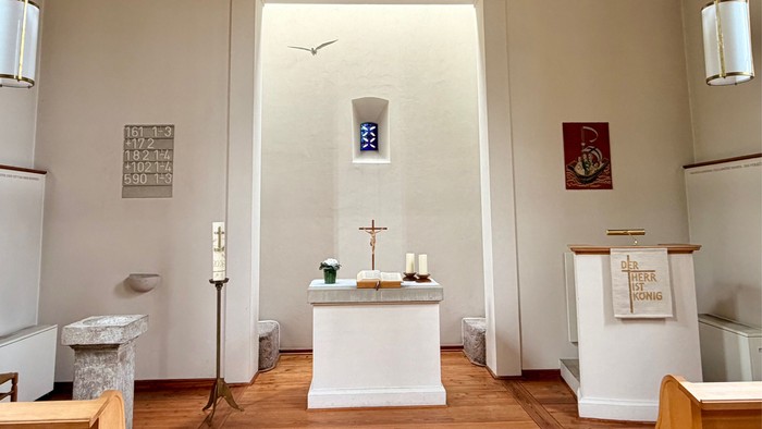 Ein ruhiger Kapelleninnenraum mit einem Altar, auf dem Kerzen stehen, einem Kreuz und religiösen Symbolen an den Wänden.