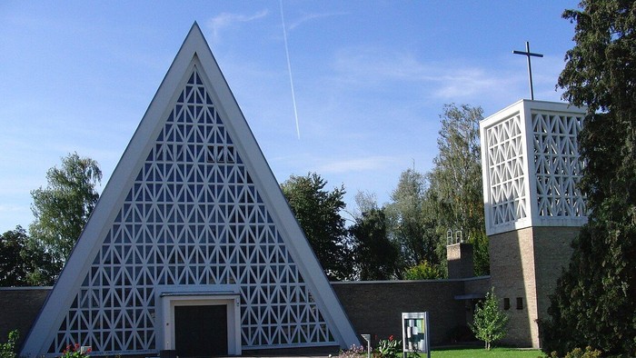 Moderne dreieckige Kirche mit geometrischem Gitterdesign und angrenzendem Glockenturm, umgeben von gestalteter Grünanlage.