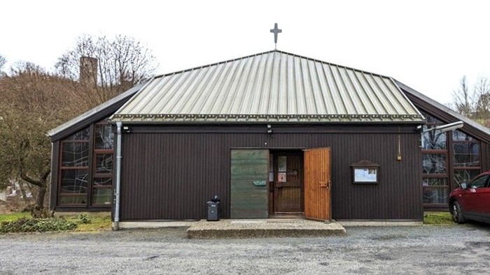 Kleine Kirchengebäude mit einem Kreuz oben drauf, geparktes Auto und schlichte Holzfassade.