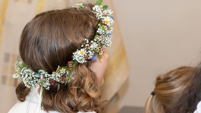 Eine Person trägt ein blumengeschmücktes Kopfteil mit lockeren Locken und ein weißes Gewand, wahrscheinlich für einen besonderen Anlass.