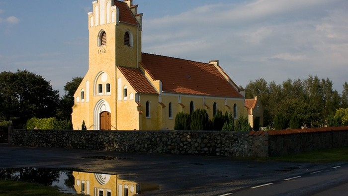 En lille gul kirke med tårn og rødt tag, omgivet af en mur og træer