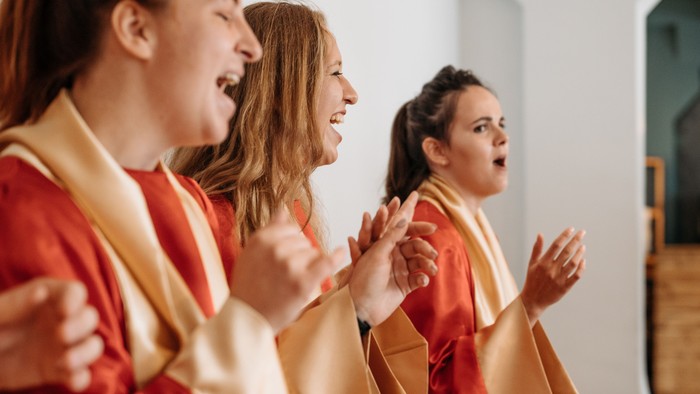 Drei Frauen in roten Chormänteln singen fröhlich in einem Kirchenambiente.