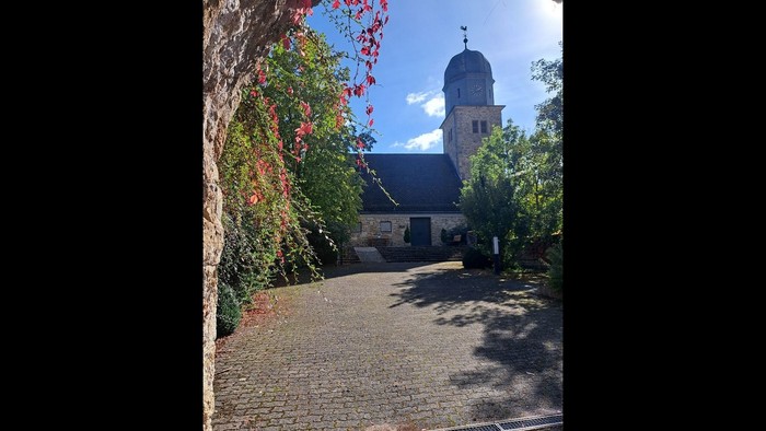 Idyllischer Kopfsteinpflasterweg, der zu einer charmanten Kirche mit hohem Glockenturm und herbstlichem Laub führt.