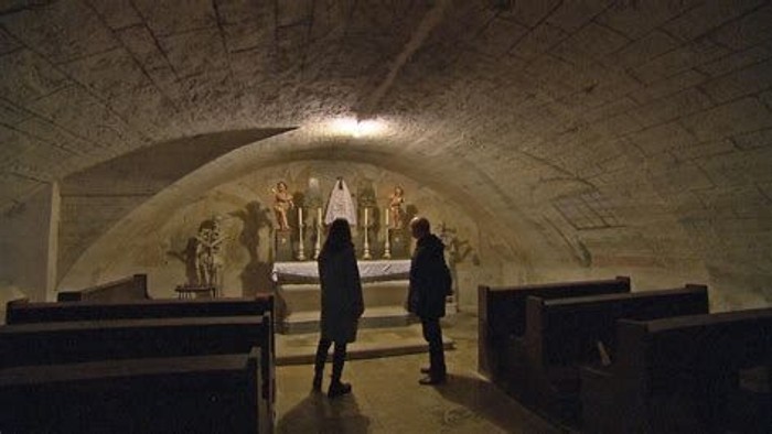 Unterirdische Kapelle mit zwei Personen, die in der Nähe eines zentralen Altars stehen, der mit religiöser Kunst geschmückt ist.