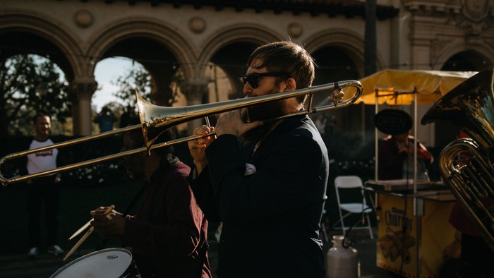 Ein Musiker spielt eine Posaune in einer Straßenkapelle mit Blechblasinstrumenten und Trommeln im Hintergrund.