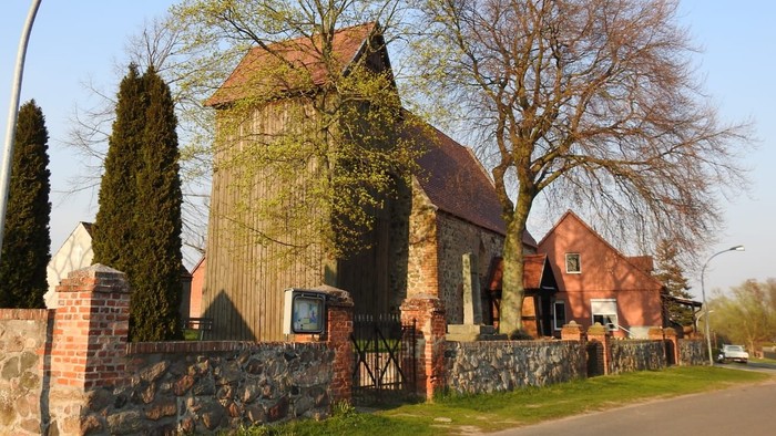 Ein rotes Backsteingebäude mit spitzem Dach und hohem Tor, umgeben von Bäumen und einem Laternenpfahl.