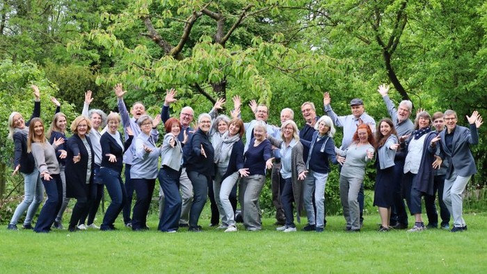Eine fröhliche Gruppe posiert freudig im Freien auf einer grasbewachsenen Wiese mit erhobenen Armen.