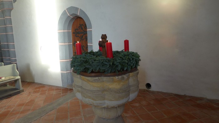 In der Kirche stehen ein steinerner Taufbrunnen mit grünem Kranz und roten Kerzen.