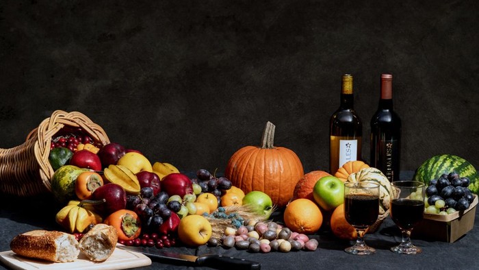 Ein Tisch mit Obstkorb, Weinflaschen und verschiedenen Früchten wie Trauben, Äpfeln und Kürbissen