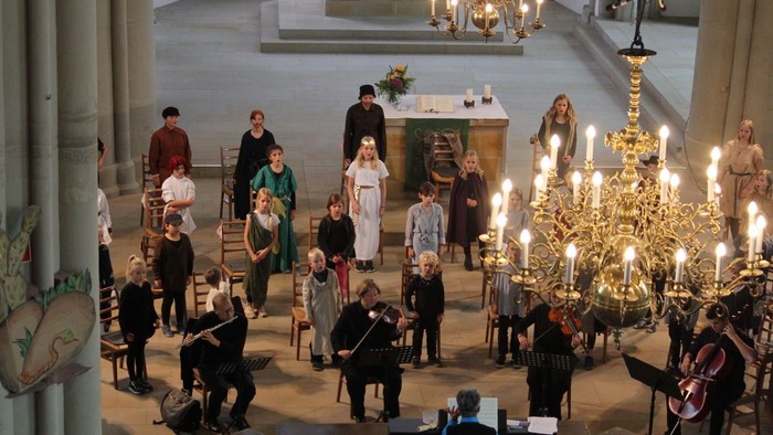 Ein Chor und Musiker treten in einer prächtigen Kirche mit kunstvollen Kerzenkronleuchtern und historischer Architektur auf.