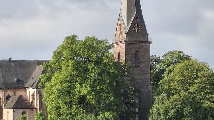 Historische Kirche mit hohem Turm und Uhr, umgeben von üppigem Grün und Wohngebäuden.
