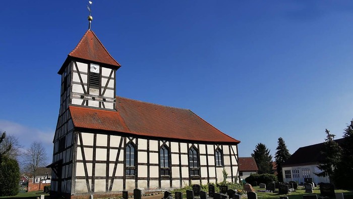 Bunte Kirche mit hohem Turm und Uhr, umgeben von Friedhof und Bäumen unter klarem Himmel. Dorfkirche Semlin