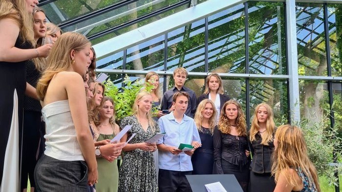 En gruppe optræder indendørs nær frodig grønne planter med en pianist, der akkompagnerer dem i et lyst og luftigt konservatorium.