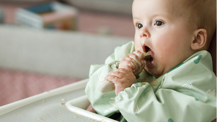 Baby isst mit einem Löffel, umgeben von verschiedenen Gläsern mit Babykost und frischem Obst.