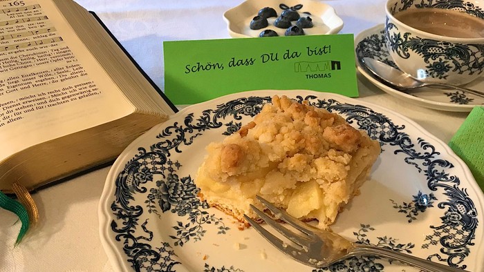 Ein Stück Kuchen auf einem dekorativen Teller neben einem aufgeschlagenen Buch und Teetassen mit einer deutschen Karte.
