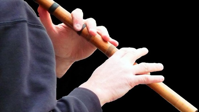Person, die eine Bambusflöte mit beiden Händen vor einem dunklen Hintergrund spielt.