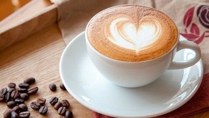 En hjerteformet latte-kunst på en kop kaffe står på en underkop omgivet af kaffebønner på en træoverflade.