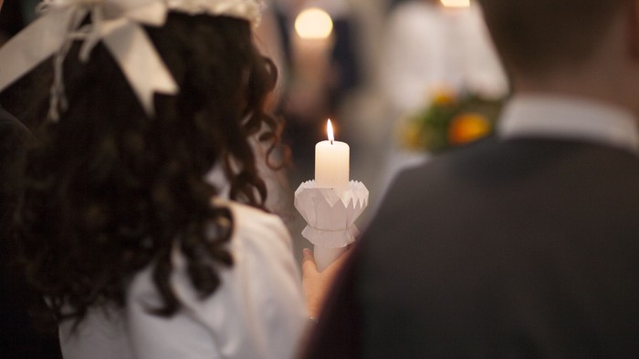 Ein Mädchen hält eine brennende Kerze in der Hand und trägt ein weißes Kleid mit Schleife.
