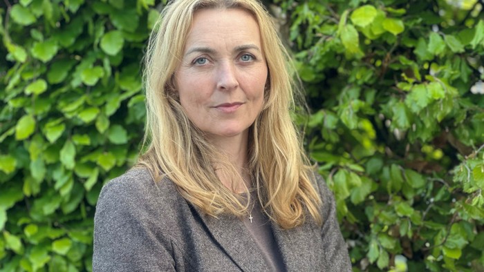 En kvinde med blondt hår i en grå blazer, der står foran en frodig grøn hæk.