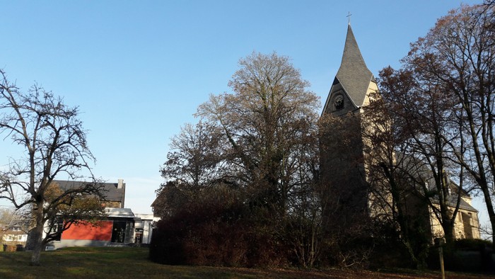 Ein Kirchengebäude mit hohem Turm, umgeben von Bäumen und Büschen, unter klarem Himmel