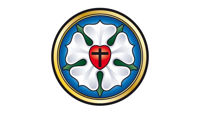 Das Bild zeigt ein Emblem mit einem roten Herz und einem Kreuz auf weißem Hintergrund, umgeben von grünen Blättern und einem blauen Ring.