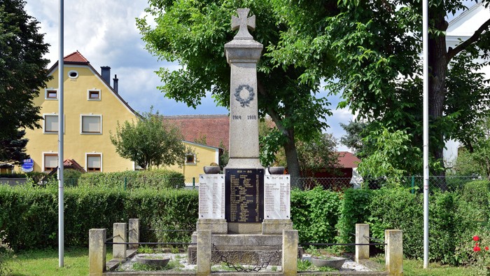 Das Bild zeigt ein Denkmal mit Uhr und Gedenktafeln vor einem gelben Gebäude.