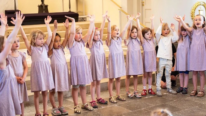 En gruppe børn i lilla tøj står med hænderne op i en kirke.