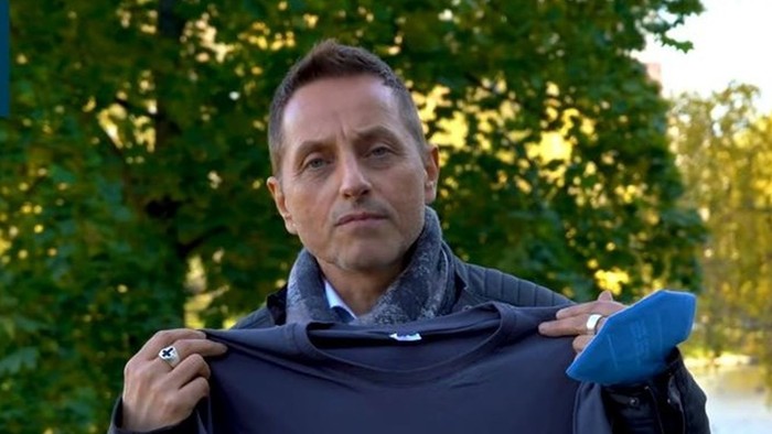 Thomas de Vachroi hält ein blaues T-Shirt, am Rand oben Text 