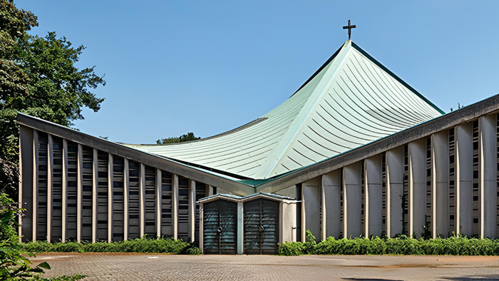 Moderne Kirche mit schlankem, geschwungenem Dach und senkrechten Betonwänden, gelegen in einer gestalteten Grünanlage.