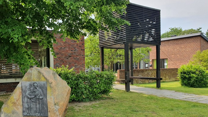 Ein ruhiger Außenbereich mit einer Steinplatte auf einem großen Felsen, umgeben von Grünflächen und modernen Pergola-Strukturen.