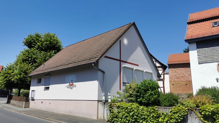 Kleine Kirche mit Kreuz an der Seite, umgeben von Büschen und Bäumen, neben einem Ziegelgebäude.