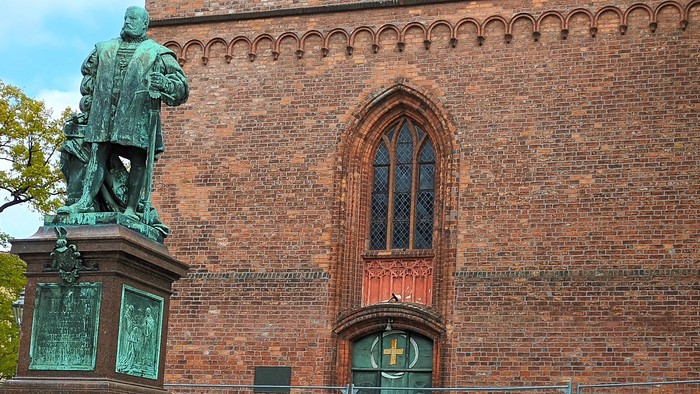 Vor einem Backsteingebäude steht eine Bronzestatue auf einem Sockel, daneben ein grüner Container und ein rotes Fahrrad.