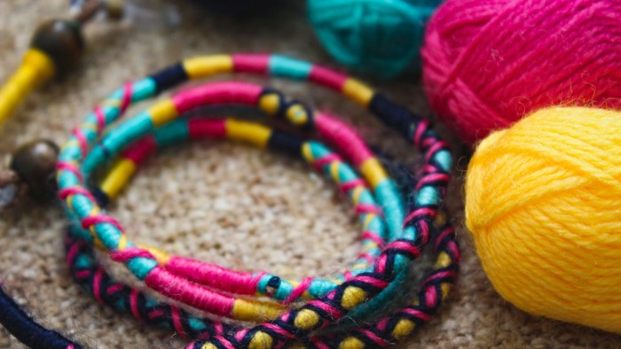 Buntes geknotetes Armband mit Perlen, umgeben von lebhaften Garnknäueln in Pink, Gelb und Türkis.