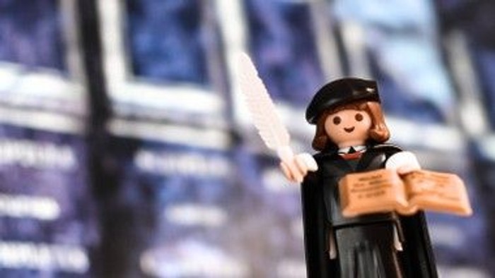 Eine Playmobil-Figur in Lehrerinnenkleidung hält ein Buch und eine Tafel.