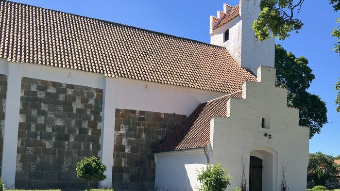 Hyggelig hvid kirke med rødt tegltag gemt i en velplejet grøn have under en klar blå himmel.