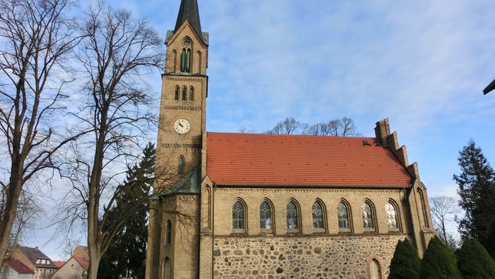 Kirche mit Uhrturm und rotem Dach, umgeben von Bäumen und Sträuchern.
