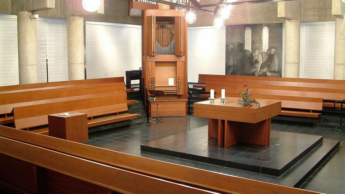 Modernes Kapelleninnenraum mit hölzernen Kirchenbänken, einem zentralen Altartisch und einer Pfeifenorgel.