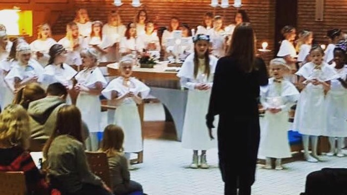 En gruppe børn i hvide tøjer synger i kirke, mens en kvinde går foran.