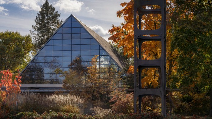 Modernes Glasgebäude, umgeben von herbstlichem Laub und einer hohen Metallskulptur.