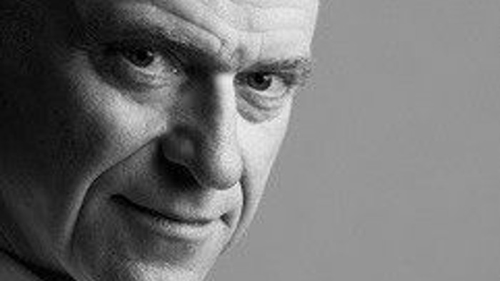 Et nærbillede i sort-hvid af en ældre mand med et let smil og begyndende skaldethed.