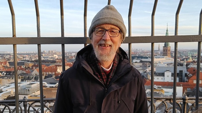 En mand i en varm frakke og hue står ved et gelænder med udsigt over en by.