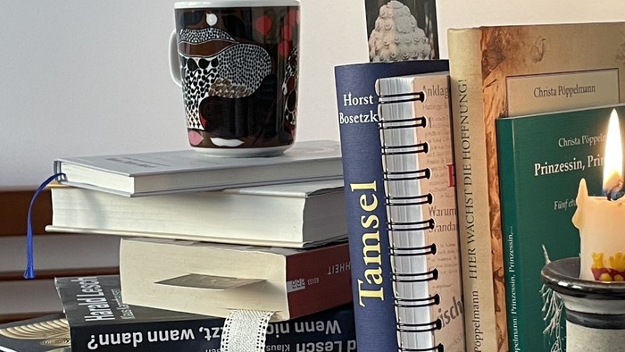 Ein gemütlicher Stapel Bücher mit einer Tasse und einer Kerze auf einem Regal, der eine Leseecken-Atmosphäre vermittelt.