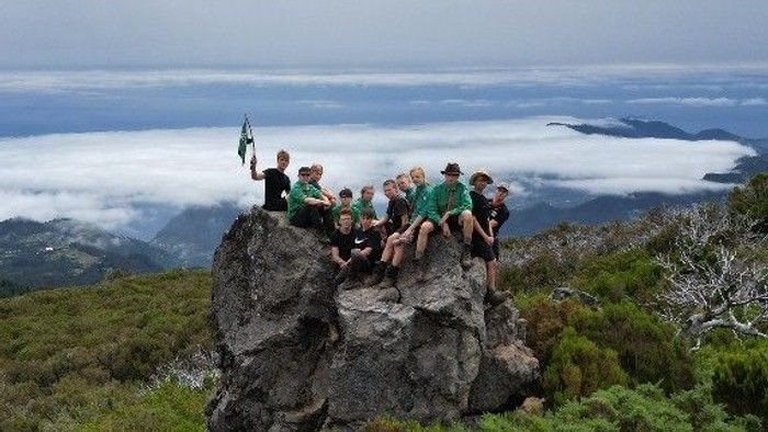 Eine Gruppe Wanderer feiert auf einem felsigen Gipfel mit einem atemberaubenden Blick auf die wolkenverhangenen Berge.