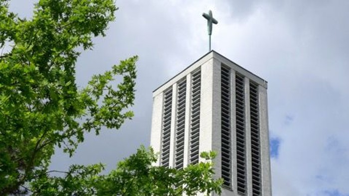 Moderner Kirchturm mit Kreuz, eingerahmt von üppigem grünem Laub unter einem bewölkten Himmel.