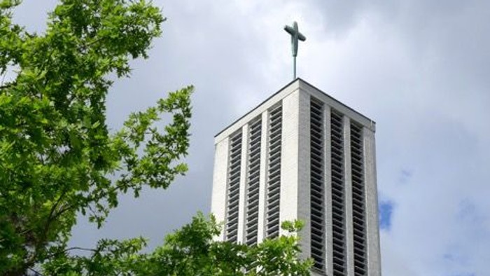Moderner Kirchturm mit Kreuz, eingerahmt von üppigem grünem Laub unter einem bewölkten Himmel.