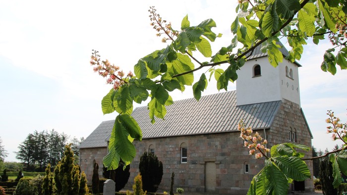 Hyggelig stenkirke med et højt klokketårn omkranset af efterårsløv og blomstrende buske.