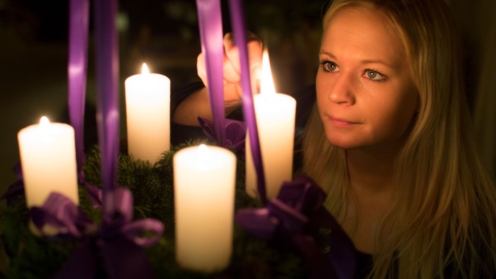 En ung pige med længe blonde hår holder en adventskranse med lysende lygter og violet bånd.